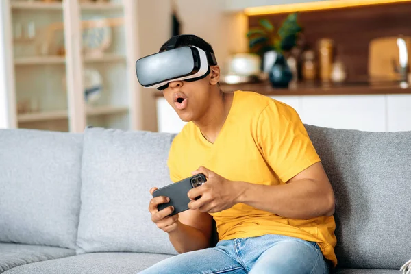 Heyecanlı karışık ırktan Latin bir adam evdeki kanepede günlük kıyafetlerle oturuyor, dijital dünyada sanal gerçeklik cihazı kullanıyor, elleriyle işaret ediyor, akıllı telefon tutuyor. Sanal dünyada oynamak