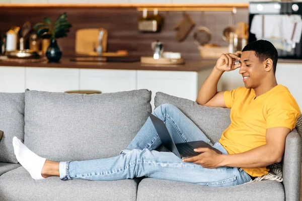 Tipo hispano atractivo positivo sentado en un sofá en casa en la sala de estar, utilizando un ordenador portátil, navegar por Internet, chatear en línea en las redes sociales, mirando a la computadora portátil, sonriendo —  Fotos de Stock