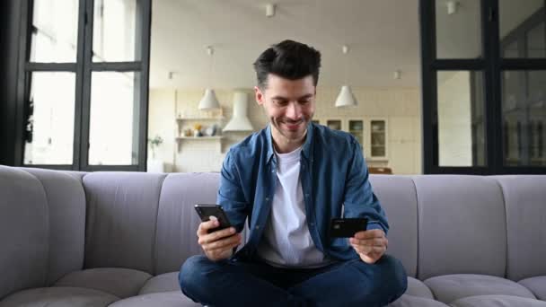 Online shopping. Happy delighted caucasian guy, sitting on couch, got big discount during checkout, uses a smartphone and credit card for payment and delivery, gestures with his hands, shouted happily — Stock Video