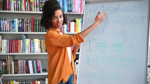 Φιλική έξυπνη νεαρή γυναίκα, αφροαμερικανή δασκάλα, στέκεται κοντά σε λευκό πίνακα, με κομψά ρούχα, διεξάγει διάλεξη ή webinar με βιντεοκλήση, gesturing με τα χέρια της, κοιτάζει κάμερα — Αρχείο Βίντεο