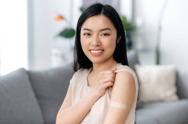 Kovid19 hastalığının tedavisi ve önlenmesi. Koronavirüs salgını ve grip sırasında aşı. Aşıdan sonra omzunda yara bandıyla tatmin olmuş Asyalı kadın gülümseyen kameraya bakıyor.