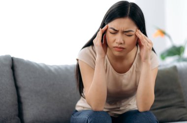 Üzgün Asyalı esmer kadın, yalnız başına kanepede oturuyor, günlük giysiler içinde, baş ağrısı çekiyor, migren oluyor, şakaklarına masaj yapıyor, gözlerini kapıyor, dinlenmeye ve tedaviye ihtiyacı var.