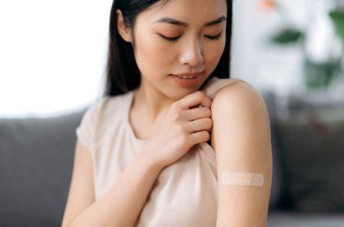 Tedavi ve önleme, aşı kavramı. Koronavirüs sırasındaki aşılar 19 salgın ve grip salgını yarattı. Aşıdan sonra omzunda yara bandıyla tatmin olmuş Asyalı kadın eline bakıyor. Yakın plan.