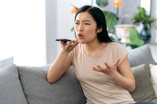 Annoyed confused asian young woman, sitting on the couch at home, holding a smartphone, irritated while talking on speakerphone or recording a voice message, dissatisfied mood