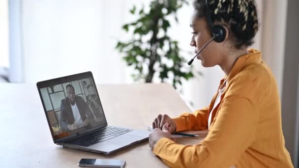 Videoconferencia en línea usando laptop. Mujer joven afroamericana en un auricular se comunica en una videollamada con su colega o cliente afroamericano, realizando una consulta en línea — Vídeos de Stock