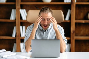 Şok olmuş Kafkasyalı genç ofis müdürü, ofiste oturuyor, şaşkınlıkla dizüstü bilgisayara bakıyor, beklenmedik bir mesaj alıyor, karlı bir iş anlaşması başarısız oluyor, duygusal yüz ifadesi.