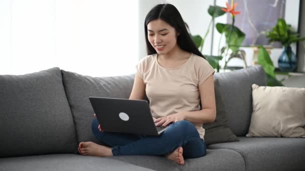 Video comunicación, conferencia en línea. Joven mujer asiática bonita, freelancer o estudiante, en ropa casual, se sienta en un sofá en casa, utiliza el ordenador portátil para comunicarse con amigos en una videollamada, charlas en línea — Vídeo de stock