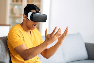 Modern, şaşkın İspanyol adam VR gözlüklerini test ediyor. Genç bir adam sanal gerçeklik gözlüğü kullanıyor, ellerine bakıyor, elleriyle jestler yapıyor, oturma odasındaki kanepede oturuyor.