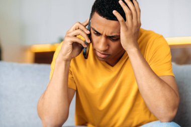 Evdeki kanepede oturan mutsuz, üzgün İspanyol genç adam, günlük sarı tişört giymiş, şaşkın, akıllı telefonla konuşuyor, kafası karışık, hoş olmayan telefon konuşmaları yapıyor.