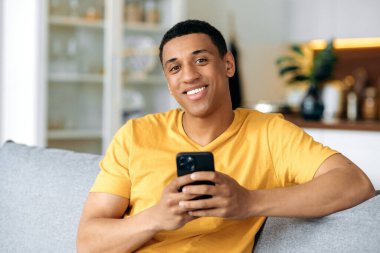 Sarı günlük tişörtlü, mutlu, İspanyol bir adam, evinde bir kanepede oturuyor, akıllı telefonunu elinde tutuyor, internette mesajlaşıyor, sosyal ağlara bakıyor, kameraya bakıyor, arkadaşça gülümsüyor.
