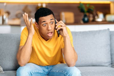 Şok olmuş, sarı tişört giymiş, akıllı telefonuyla konuşan, kanepede oturan İspanyol şaşkın genç adam, ezici haberler aldı, şaşkın yüz ifadesi, el hareketi