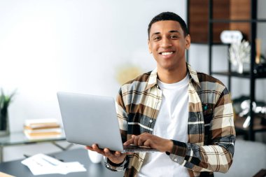 Yakışıklı, karizmatik, modern İspanyol bir adam şık bir gömlek giymiş, ofis çalışanı, serbest çalışan ya da öğrenci, elinde dizüstü bilgisayar, masanın yanında duruyor, kameraya bakıyor, arkadaşça gülümsüyor.