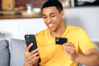 Neşeli, pozitif İspanyol asıllı genç adam oturma odasında, sarı tişört giymiş, akıllı telefon ve kredi kartını online alışveriş için kullanıyor, ödeme için veri giriyor, çevrimiçi işlem, gülümsüyor.