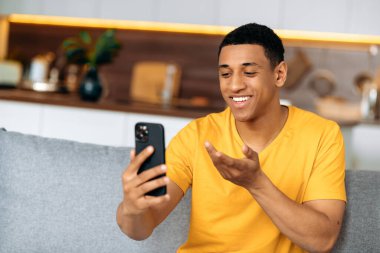 Akıllı telefon kullanarak çevrimiçi video iletişimi. Pozitif ispanyol bir adam oturma odasında bir kanepeye oturur, akıllı bir telefon tutar, video görüşmesinde konuşur, el kol hareketi yapar, mutlu bir şekilde gülümser.