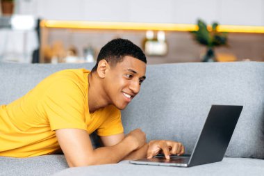 İspanyol asıllı, sarı tişörtlü, tembel bir hafta sonu geçiriyor, kanepede yatıyor, dizüstü bilgisayar kullanıyor, sosyal ağlarda arkadaşlarıyla sohbet ediyor, haberleri izliyor, internetten eve sipariş veriyor, gülümsüyor.