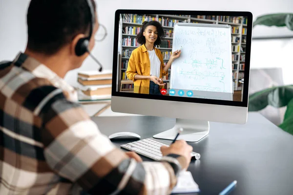 Conceito de e-learning, webinar, palestra online. Sobre a visão do ombro do estudante masculino em uma tela de computador, professor americano africano amigável fêmea na tela conduz uma lição on-line, um cara toma notas — Fotografia de Stock