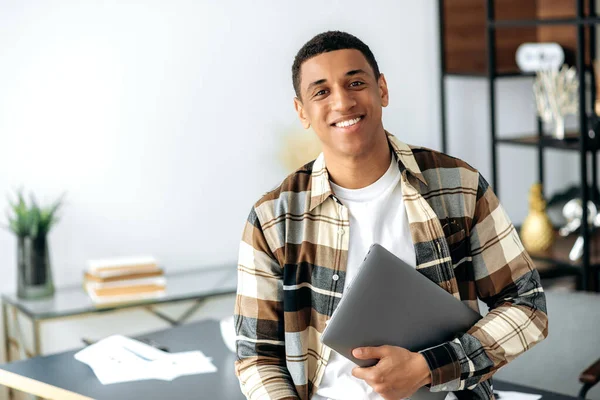 Retrato de un guapo tipo hispano moderno y carismático, vestido con camisa elegante, oficinista, freelancer o estudiante, sosteniendo un portátil, parado cerca de una mesa, mirando a la cámara, sonriendo amigable — Foto de Stock