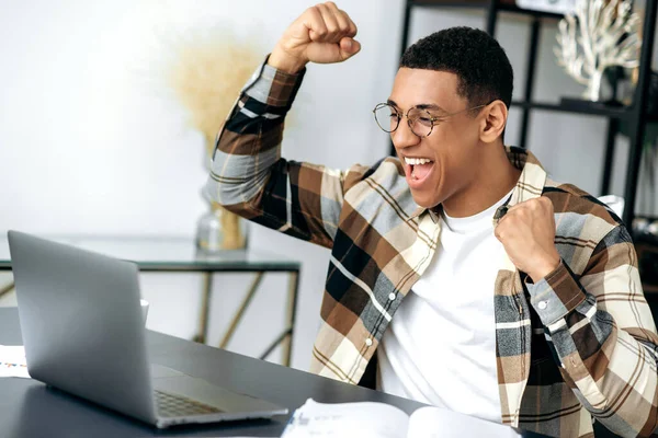 Feliz chico hispano feliz y exitoso con gafas, vestido con estilo, empleado, freelancer o estudiante se sienta en el escritorio, haciendo gestos con sus manos regocijándose en la victoria, el éxito, buen negocio, grandes ganancias —  Fotos de Stock