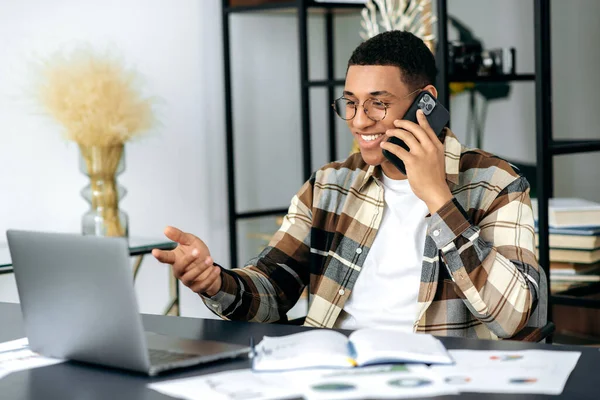 Ocupado alegre hispânico elegante jovem, trabalhador de escritório ou freelancer, sentado na mesa de trabalho, tendo uma conversa agradável telefone celular durante o tempo de trabalho, sorrindo amigável, ter um bom humor — Fotografia de Stock
