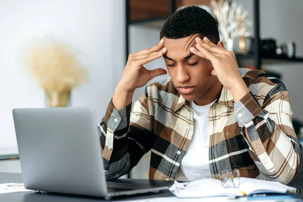 Joven latino deprimido y cansado, con ropa casual y elegante, freelancer, sentado en su escritorio, masajeando sus sienes con los ojos cerrados, pensando en un proyecto, tiene dolor de cabeza, migraña, exceso de trabajo —  Fotos de Stock