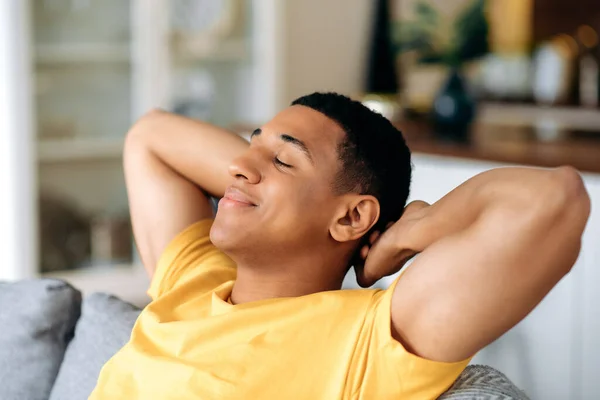 Relaxado satisfeito calma hispânico cara em uma camiseta amarela, descansando no sofá em casa com os olhos fechados, jogando as mãos atrás de sua cabeça, sonhando com umas férias, descansando do trabalho, sorrindo — Fotografia de Stock