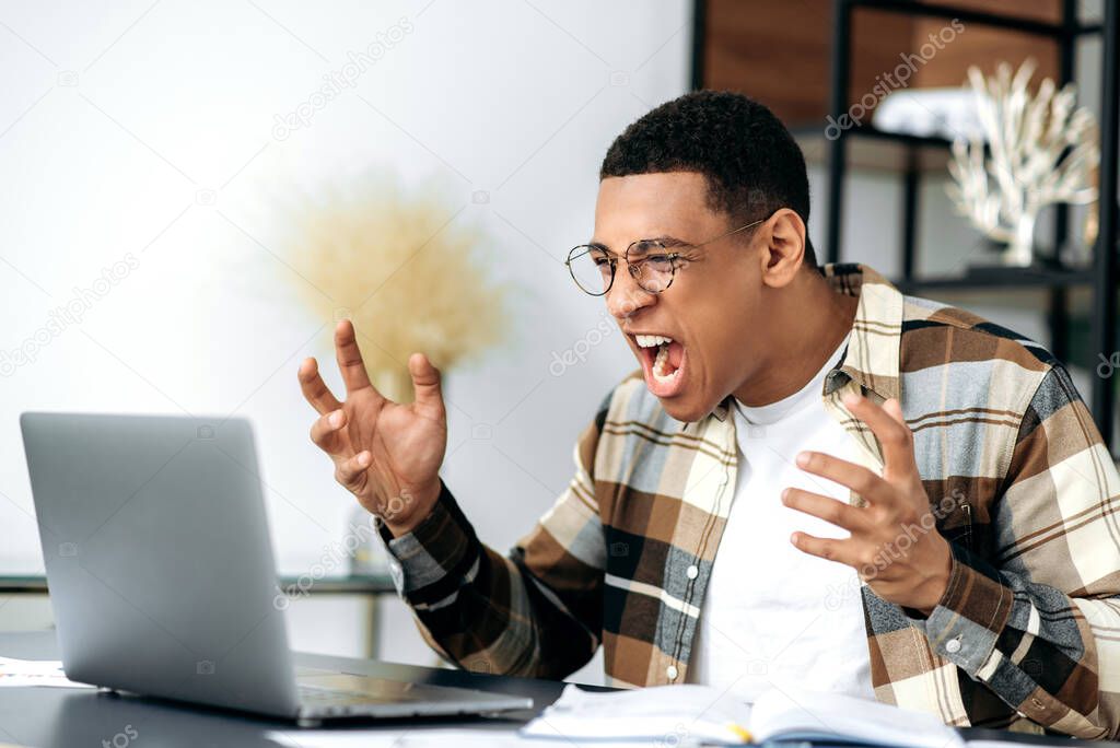 Angry annoyed hispanic young man with glasses, freelancer, manager or student, emotionally yells at a laptop, got bad news, failed, lost, gesturing with hands, very irritated and frustrated