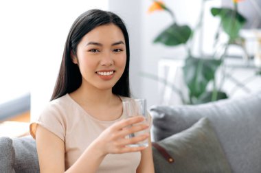 Sağlıklı yaşam tarzı kavramı. Pozitif, güzel Çinli ya da tişörtlü Japon genç bir kadın bir bardak temiz su tutuyor, kameraya bakıyor, gülümsüyor, sağlığına önem veriyor, günlük su normlarını içiyor.