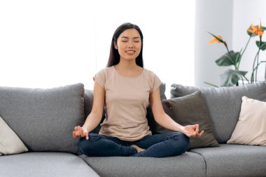 Meditasyon, rahatlama, evde tembellik. Güzel, sakin Asyalı, günlük kıyafetler giymiş genç bir kadın nilüfer çiçekli koltukta gözleri kapalı oturur, gülümser, meditasyon yapar, düşüncelerini boşaltır, sakinlik kavramı oluşturur.