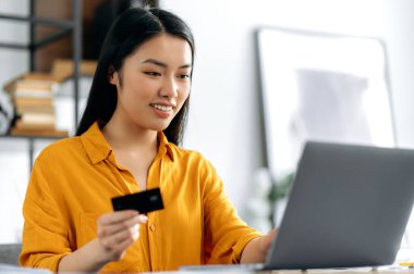 İnternetten alışveriş. Pozitif genç Asyalı esmer kadın bir masada oturuyor, online alımları ve teslimatları ödemek için dizüstü bilgisayar ve kredi kartı kullanıyor, ayrıntılara giriyor, karta bakıyor, gülümsüyor. Çevrimiçi aktarım