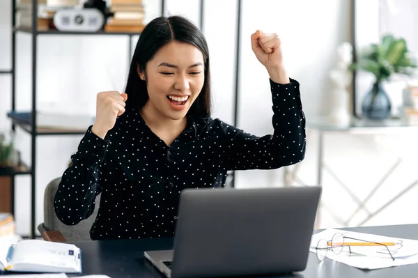 Happy positif wanita muda asia yang sukses, berpakaian rapi, pekerja kantor, pekerja lepas atau mahasiswa duduk di meja kerja, memberi isyarat dengan tangannya bersukacita dalam kemenangan, sukses, kesepakatan yang bagus, keuntungan besar — Stok Foto