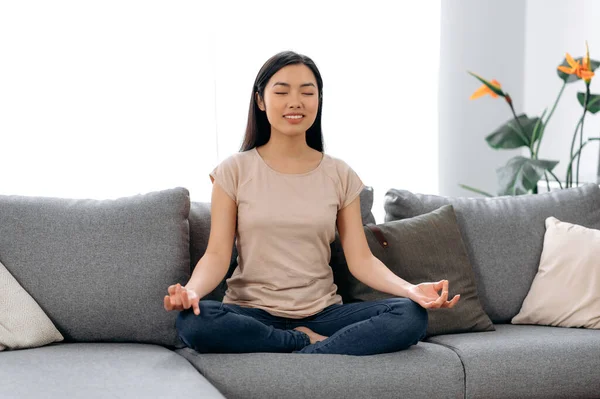 Meditasyon, rahatlama, evde tembellik. Güzel, sakin Asyalı, günlük kıyafetler giymiş genç bir kadın nilüfer çiçekli koltukta gözleri kapalı oturur, gülümser, meditasyon yapar, düşüncelerini boşaltır, sakinlik kavramı oluşturur.