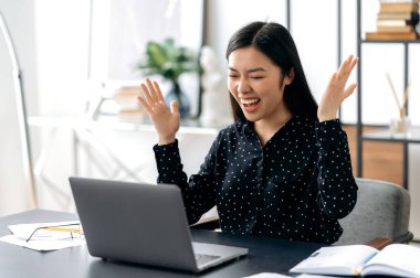 Neşeli, başarılı Asyalı genç kadın, şık giyimli, ofis çalışanı, serbest çalışan ya da öğrenci çalışma masasında oturuyor, elleri zaferle, başarıyla, iyi anlaşmayla, büyük kazançla el kol geziyor.