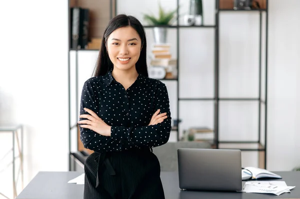 Portrét úspěšné sebevědomé hezké, mladé asijské obchodní žena nebo na volné noze stojí u pracovního stolu v kanceláři, na sobě formální stylové oblečení, paže zkřížené, dívá se do kamery, usmívá přátelsky — Stock fotografie