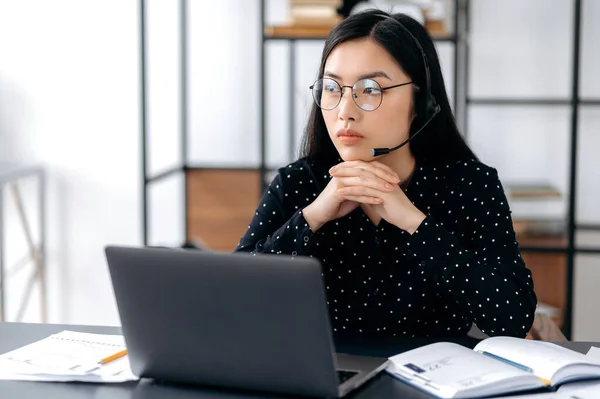 Verontrust aziatische jonge brunette vrouw met bril en een headset, een support operator of manager, zitten aan een bureau, kijken bedachtzaam opzij, ongelukkig, ervaren van stress, moet rusten, bezorgd — Stockfoto