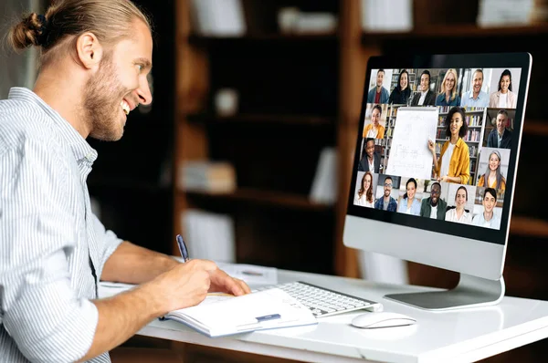 Online oktatás. Okos kaukázusi fickó tudást szerez online tanítás közben. A fiatalember jegyzetel a webinárium alatt, videóhívásról szóló előadást hallgat. Női tanár és a diákok a képernyőn — Stock Fotó