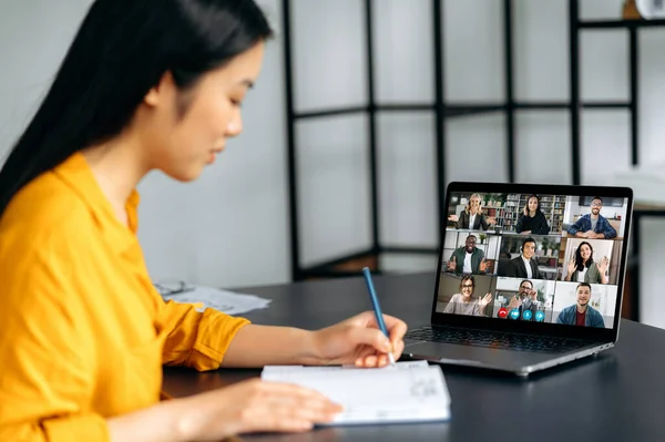 Una estudiante asiática inteligente gana conocimiento, escucha un seminario en línea, toma notas durante una conferencia de video, aprendiendo de forma remota. Entrenador y diferentes personas en la pantalla del portátil.Videollamada —  Fotos de Stock