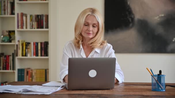 Очаровательная белая деловая женщина средних лет в белой рубашке сидит за рабочим столом, элегантная блондинка задумчиво смотрит в сторону, думает о стратегии проекта, работает с ноутбуком, делает заметки — стоковое видео