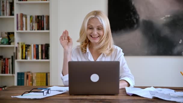 Καυκάσιος ελκυστική κομψή ενήλικη επιχειρηματίας σε ένα κομψό λευκό πουκάμισο κάθεται σε ένα χώρο εργασίας, χρησιμοποιεί φορητό υπολογιστή για την επικοινωνία με τους συναδέλφους με βιντεοδιάσκεψη, χαιρετισμό συνομιλητή, φιλικό χαμόγελο — Αρχείο Βίντεο