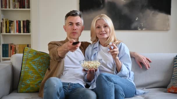 Ver una película juntos. Mediana edad feliz casada pareja caucásica sentado en casa en el sofá, viendo la televisión, comer palomitas de maíz, ver películas favoritas, pasar tiempo libre en casa, felices juntos, sonreír — Vídeo de stock