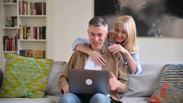幸せな白人の喜びの配偶者はソファの上で自宅で時間を過ごし、友人や家族とのオンライン通信にラップトップを使用してビデオ会議で挨拶し、手を振って笑顔で挨拶します。ビデオ通話のコンセプト — ストック動画
