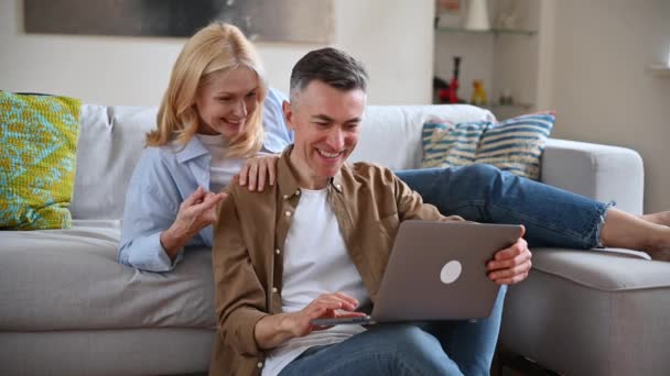 Gelukkig Kaukasisch familie paar, volwassen vrouw en echtgenoot thuis op een bank, in casual kleding, met behulp van laptop, surfen op internet, sociale netwerken, chatten met vrienden of familie, op zoek naar huishoudelijke artikelen — Stockvideo