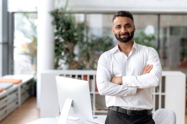Resmi bir erkek portresi. Beyaz gömlekli başarılı Hintli işadamı ya da menajer ofiste çalışma masasının yanında duruyor, kollarını kavuşturup doğrudan kameraya bakıyor ve dostça gülümsüyor.