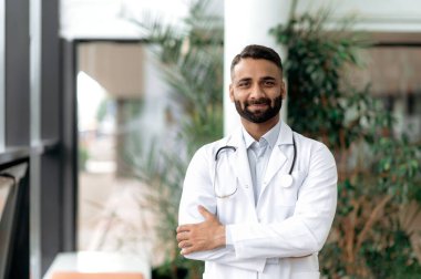 Klinikte kolları çapraz duran, boynunda steteskop ile tıbbi giysiler giymiş kameraya bakarak dostça gülümseyen Hintli bir tıp doktorunun portresi. Tıbbi uzman