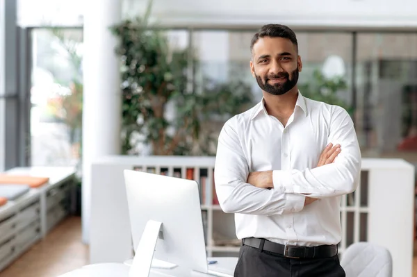 Retrato formal de hombre de negocios. Confiado empresario o gerente indio exitoso, en camisa blanca, está cerca de su escritorio en la oficina, los brazos cruzados, mira directamente a la cámara y sonríe amigable — Foto de Stock