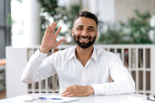 Indiano amigável inteligente homem positivo, ceo ou gerente, senta-se na mesa de trabalho, conversando com colegas através de videoconferência, interlocutor saudação com gesto de mão, sorrisos, ter conversa on-line — Fotografia de Stock