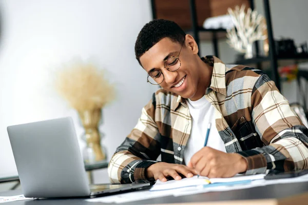 Joven latino inteligente y positivo con gafas, sentado a la mesa, usando un portátil, tomando notas, estudiando o trabajando en línea, escuchando un webinar, adquiriendo conocimiento, sonriendo —  Fotos de Stock