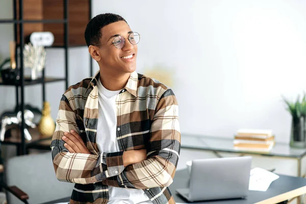 Retrato de un atractivo joven latino de raza mixta positivo y seguro con gafas, diseñador o freelancer, vestido con estilo casual, parado cerca de un escritorio con los brazos cruzados, mirando hacia un lado, sonríe —  Fotos de Stock