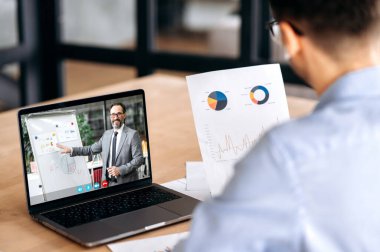 Finansal grafikler tutan beyaz bir erkek yöneticinin omuz hizasında, olgun başarılı CEO şirketiyle çevrimiçi video konferansı yaparken, dizüstü bilgisayar kullanarak, ofiste otururken, konsantre olarak,