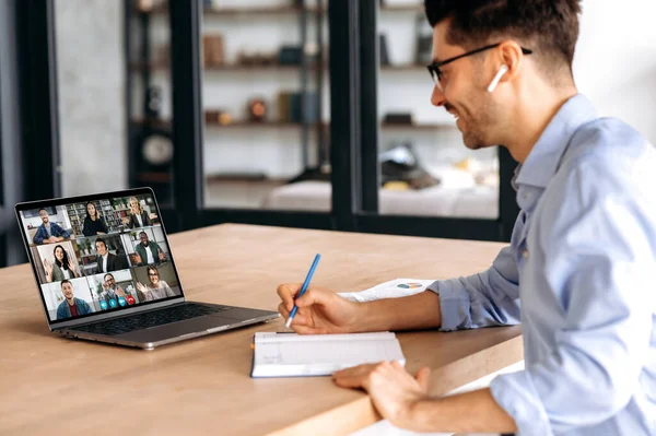 Side view to a confident male manager, taking notes during online business meeting with multinational colleagues, using laptop, sitting in office, planning marketing strategy, listening colleorkers — стоковое фото