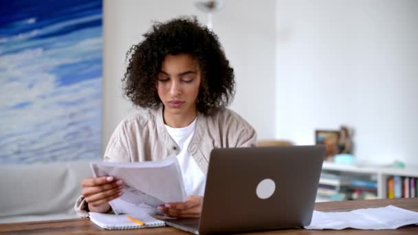 Έξυπνη εστιασμένη αυτοπεποίθηση σγουρά μαλλιά αφροαμερικανή νεαρή γυναίκα σε casual ρούχα, ελεύθερος επαγγελματίας ή φοιτητής, κάθεται στο σαλόνι στο φορητό υπολογιστή, εργασίας ή μελέτης εξ αποστάσεως, αναλύοντας οικονομικά έγγραφα — Αρχείο Βίντεο